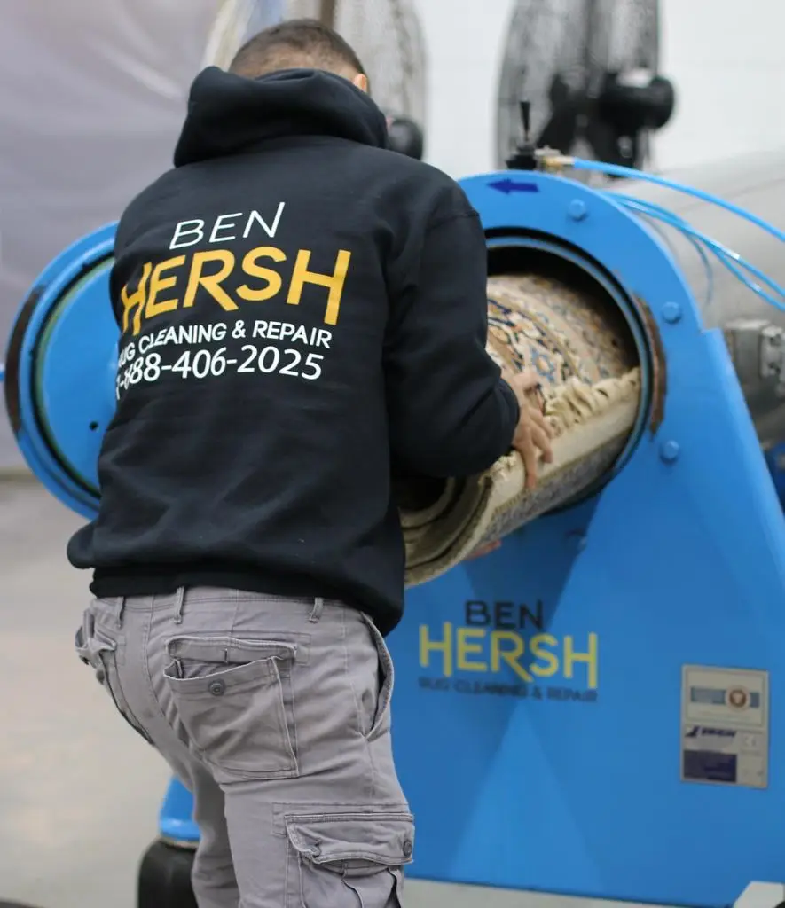 A person wearing a black hoodie that reads Ben Hersh Rug Cleaning & Repair 888-406-2025 is operating a large blue machine designed for area rug cleaning. The machine also displays the same company name and contact information. Various tools and industrial fans are visible in the background.