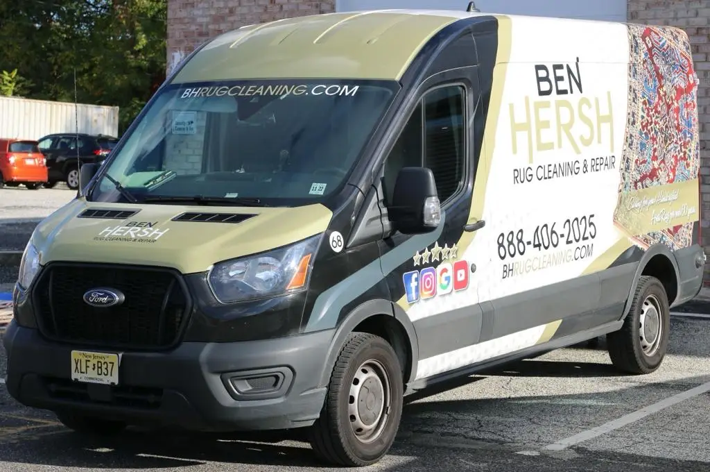 A van with Ben Hersh Rug Cleaning & Repair signage is parked in a lot, showcasing contact info and social media icons. Prominently black and white, it features a vibrant rug pattern. Known for being among the best rug cleaning companies near me, its license plate reads XLF-B37 amid a scene of buildings and cars.