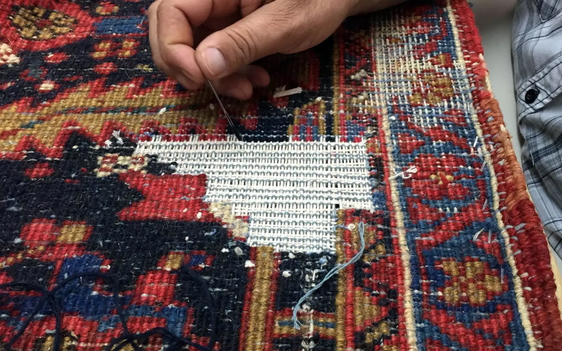 A person is meticulously repairing a colorful woven rug with a needle and thread. The rug, boasting intricate red, blue, and yellow patterns, features a large white patch being sewn in the middle. For pristine results similar to this restoration, consider consulting the best rug cleaning companies near you.