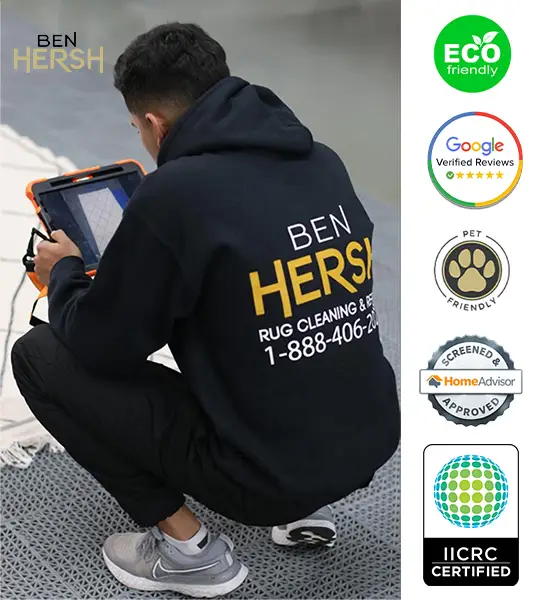 A person in a black hoodie with Ben Hersh Rug Cleaning & Repair printed on it is kneeling and cleaning a rug. Showcasing badges for eco-friendliness, Google verified reviews, pet friendliness, HomeAdvisor approval, and IICRC certification, it's clear they're among the best rug cleaning companies near me.