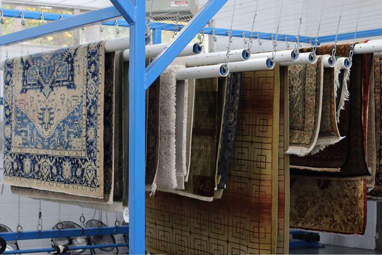 Rugs in various colors and patterns, including blue and beige designs, hang from blue metal racks in a facility known for oriental rug cleaning. The rugs are suspended on white cylindrical rods with plastic hooks against a backdrop of a white tiled wall.