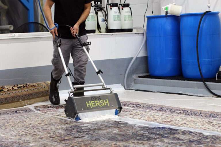 A person is operating a BEN HERSH carpet cleaning machine on an oriental rug, creating suds as it works. In the background, labeled containers line the shelf and two large blue barrels sit on the floor. The setting feels industrious and clean—ideal for the best rug cleaning companies near me.