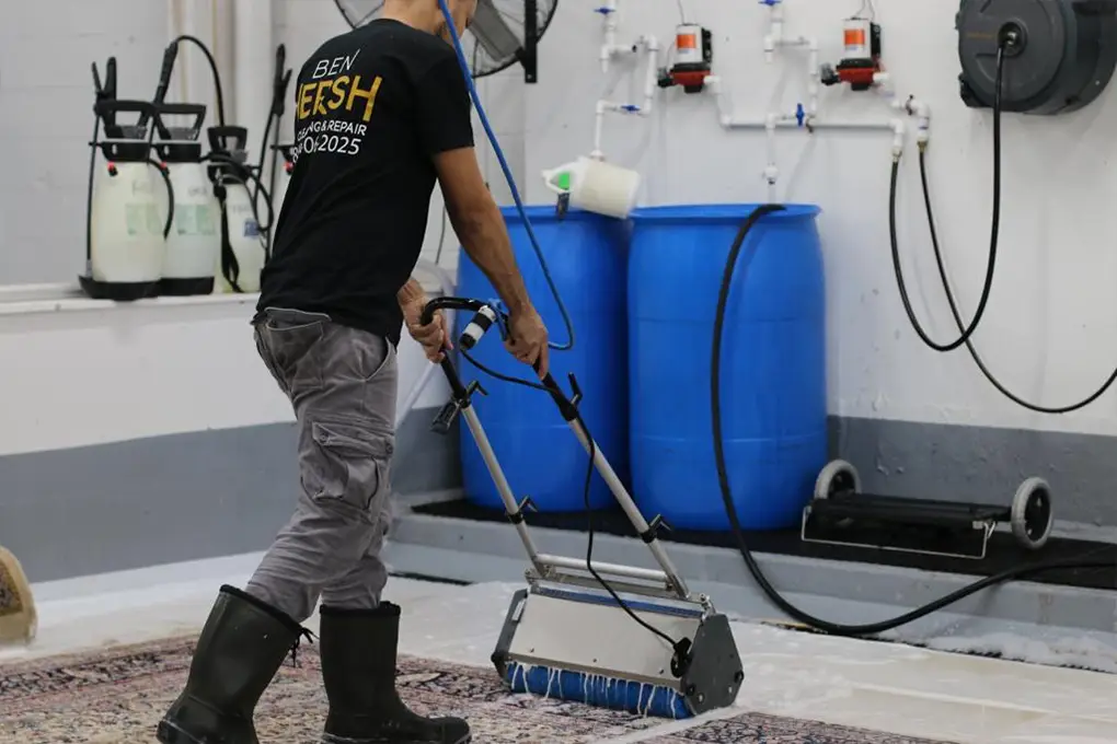 A person operates a carpet cleaning machine over a patterned rug in a workshop. Sporting black boots, gray pants, and a T-shirt with text on the back, they expertly navigate the room, which houses two large blue barrels and supplies—a scene reminiscent of top area rug cleaning near me services.