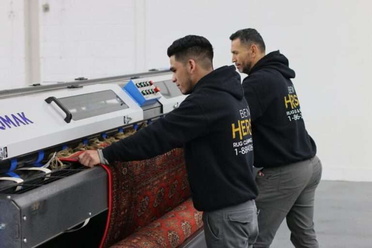 Two men in black hoodies operate a rug cleaning machine indoors, expertly restoring an oriental rug. The men focus intently on the patterned red fabric as their hoodies display a business logo and contact number. In the bright room with white walls and a gray floor, they embody one of the best rug cleaning companies around.