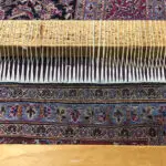 A wooden weaving loom displays strings stretched vertically, showcasing a partially completed intricate Persian rug with detailed blue, red, and white patterns. The floor beneath is wooden, with another similar rug in the background. Find the best rug cleaning companies near me to maintain such beauty.