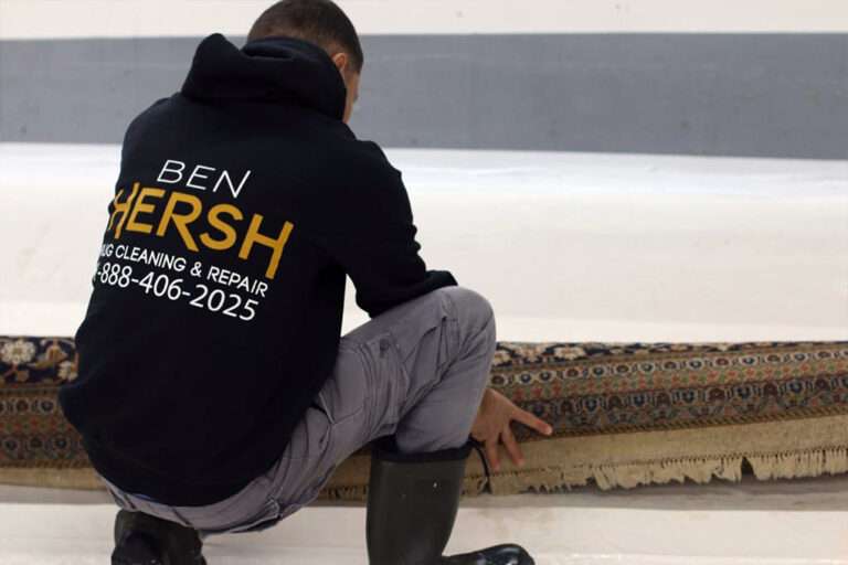 A person wearing gray pants and black boots is examining a rolled-up carpet with intricate patterns. Sporting a black hoodie that reads BEN HERSH RUG CLEANING & REPAIR with a phone number, they seem to know the best rug cleaning companies near me. The background is a neutral-colored wall.