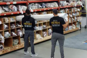 Two men wearing black shirts with BEN HERSH carry a rolled-up carpet in a warehouse, where shelves brim with various carpets wrapped in plastic. The gray flooring adds to the organized ambiance, showcasing a selection akin to what best rug cleaning companies might handle.