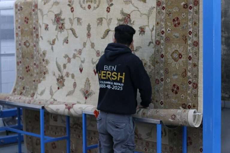 A person wearing a black hoodie with BEN HERSH CLEANING & REPAIR and a phone number on the back is inspecting an oriental rug on a blue metal frame. The intricately patterned rug, featuring floral designs in beige, red, and green tones, showcases expertise from one of the best rug cleaning companies near you.