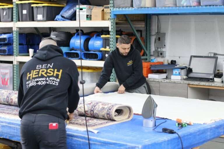 In a workshop, two people focus on a rolled-out rug atop a blue table. One measures the fabric while the other inspects it. The person on the left sports a black hoodie with Ben Hersh Rug Cleaning & Repair emblazoned on the back—one of the best rug cleaning companies near me. An iron and assorted tools lie in the background.
