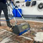 Operating a Ben Hersh carpet cleaning machine, the individual in black boots and dark pants maneuvers over a patterned carpet with soap suds. The backdrop features equipment and hoses on the wall, reminiscent of top rug cleaners in New Jersey.