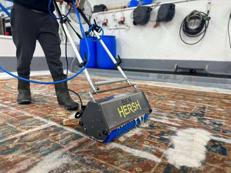 Operating a Ben Hersh carpet cleaning machine, the individual in black boots and dark pants maneuvers over a patterned carpet with soap suds. The backdrop features equipment and hoses on the wall, reminiscent of top rug cleaners in New Jersey.