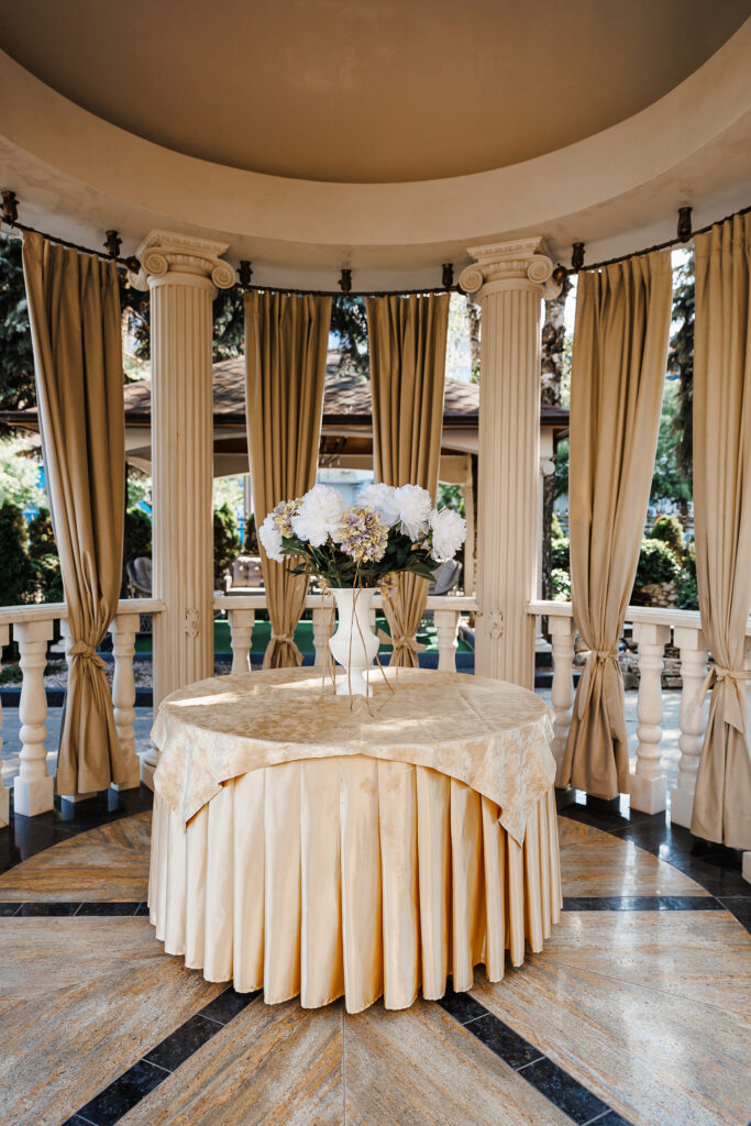 Rug Cleaning - A round table draped in gold cloth sits at the center of a circular pavilion with pristine white columns and beige curtains, impeccably maintained with drape cleaning. The table cradles a white vase brimming with white flowers. The open pavilion reveals trees beyond, while the floor boasts a geometric tiled pattern.