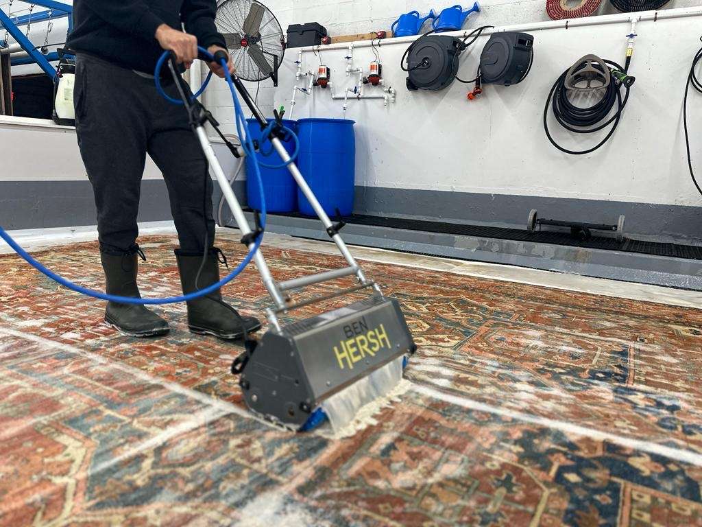 A person in black clothes and rubber boots expertly operates a carpet cleaning machine labeled Ben Hersh on a patterned rug. Soap suds cover the wet floor, surrounded by cleaning equipment, a blue drum, hoses, and a large fan mounted on the white wall.