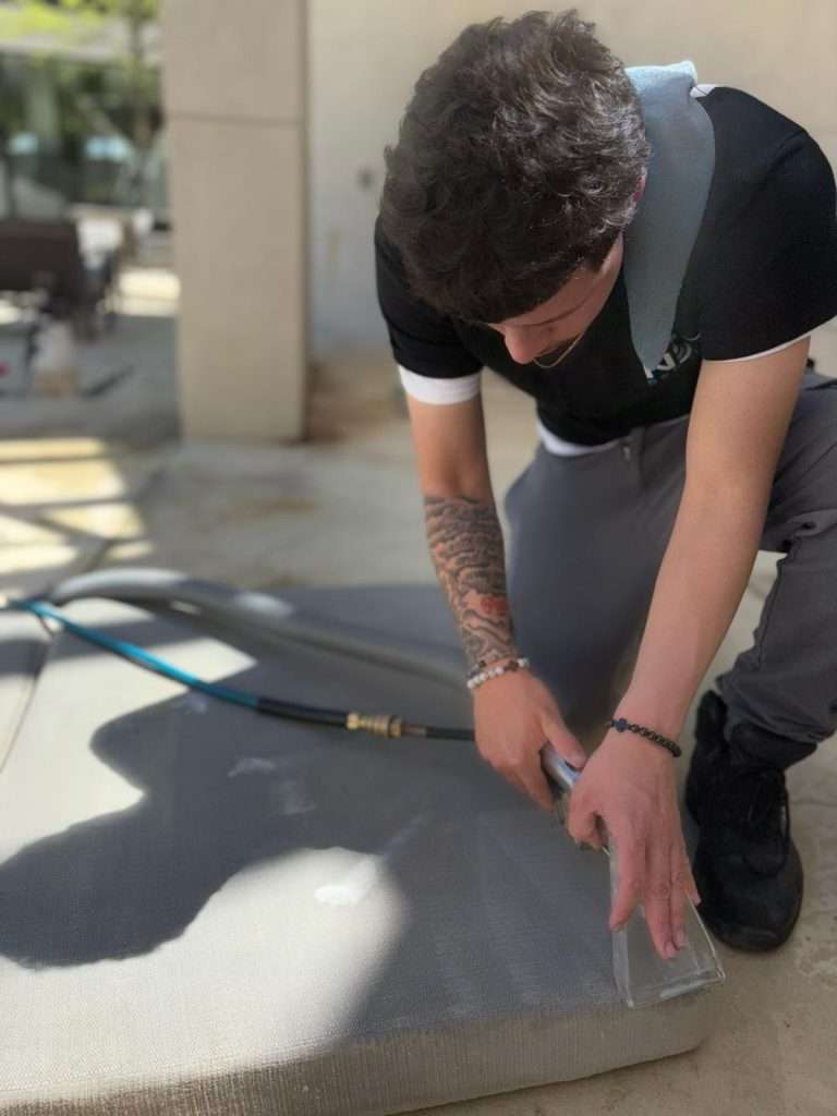 A young man with a tattooed right arm is crouched down outdoors, using a utility knife to cut a large piece of gray mesh material—possibly for upholstery cleaning. A blue hose lays on the concrete nearby, sunlight and building shadows in the background.