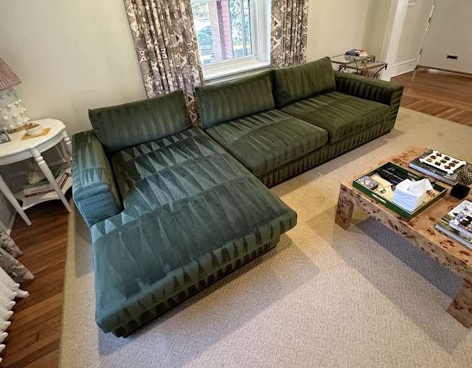 A green velvet sectional sofa with a chaise lounge sits on a beige carpet. Nearby, a white round side table with books and a lamp, patterned curtains, and a square coffee table holding books and tissues create the ideal spot for Sofa Cleaning in this cozy living room.