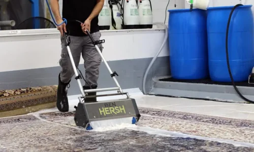 A person is using a Ben Hersh machine to clean an oriental rug with soapy water. Blue barrels and white containers with nozzles, likely for cleaning supplies, stand against the gray and white wall. For expert rug cleaners in New Jersey, this scene epitomizes efficiency and care.