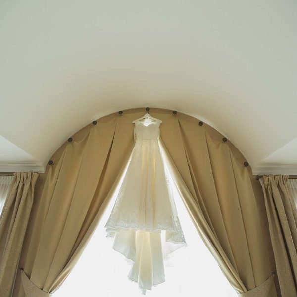 Rug Cleaning - A white wedding dress hangs elegantly between two golden curtains under an arched window. The room is brightly lit, showcasing the best in delicate lace and flowing fabric of the gown. Soft natural light filters through sheer curtains, creating a serene and romantic atmosphere.