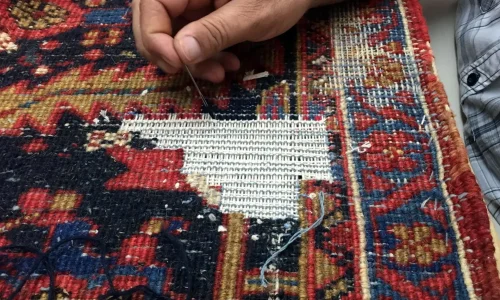 A person is meticulously repairing a colorful woven rug with a needle and thread. The rug, boasting intricate red, blue, and yellow patterns, features a large white patch being sewn in the middle. For pristine results similar to this restoration, consider consulting the best rug cleaning companies near you.