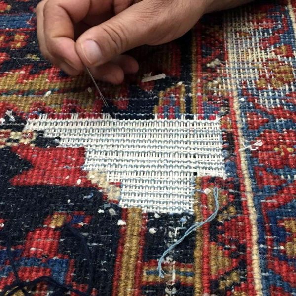 A person is meticulously repairing a colorful woven rug with a needle and thread. The rug, boasting intricate red, blue, and yellow patterns, features a large white patch being sewn in the middle. For pristine results similar to this restoration, consider consulting the best rug cleaning companies near you.