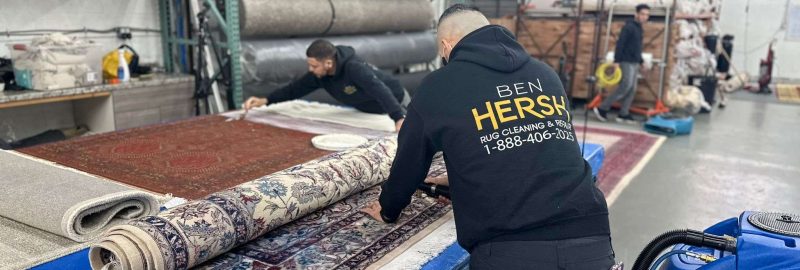 Two men are diligently cleaning and rolling rugs in a well-organized workshop. One wears a black hoodie with Ben Hersh and a phone number on the back. In the foreground, a blue cleaning machine from one of the best rug cleaning companies near me stands ready for use amidst neatly stored rugs.