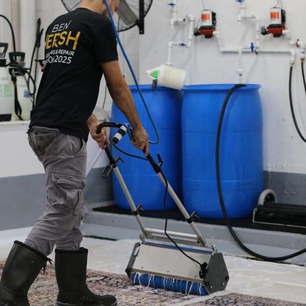 A person operates a carpet cleaning machine over a patterned rug in a workshop. Sporting black boots, gray pants, and a T-shirt with text on the back, they expertly navigate the room, which houses two large blue barrels and supplies—a scene reminiscent of top area rug cleaning near me services.