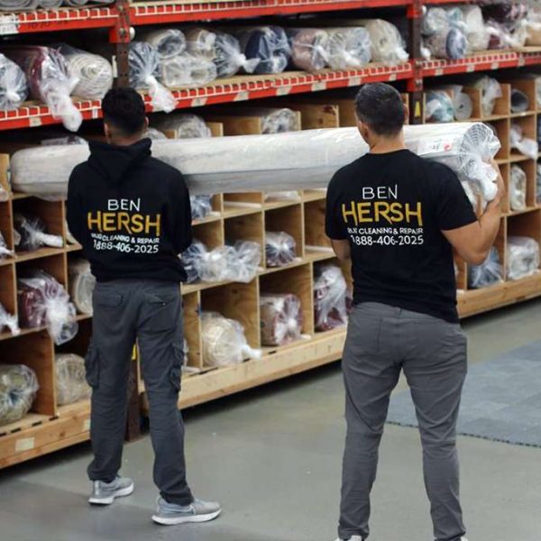 Two men wearing black shirts with BEN HERSH carry a rolled-up carpet in a warehouse, where shelves brim with various carpets wrapped in plastic. The gray flooring adds to the organized ambiance, showcasing a selection akin to what best rug cleaning companies might handle.